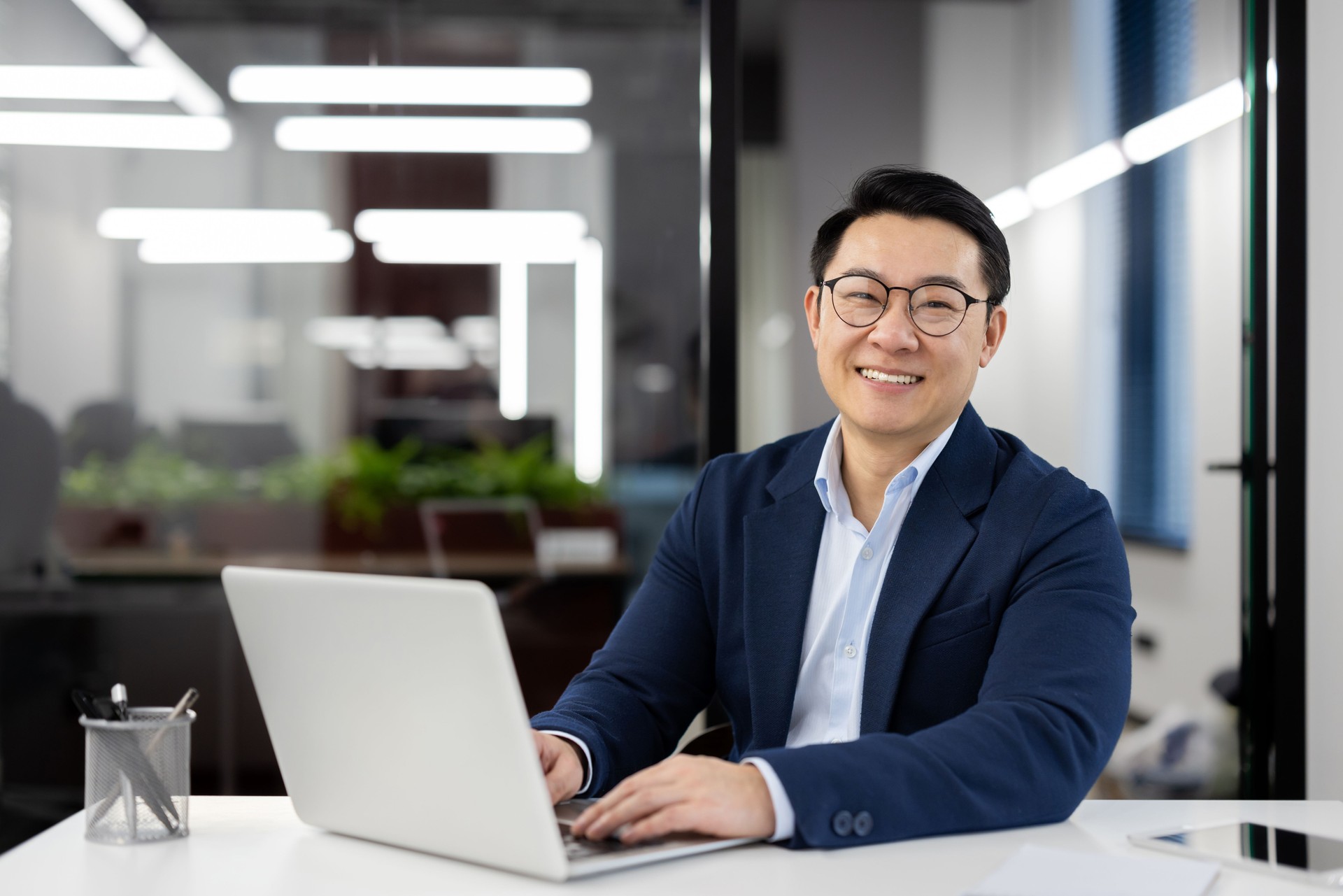 Portrait of successful mature asian businessman, experienced boss in business suit smiling and looking at camera, financier investor inside office at workplace working with laptop, satisfied man